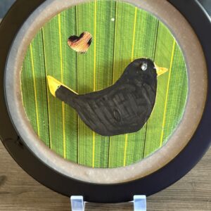 A round, framed bird is mounted on a green, striped background resembling a leaf. The black bird is accented with a yellow beak and yellow tail and a gemstone is used for the bird’s eye. Above the bird, there is a small yellow and brown heart shape.