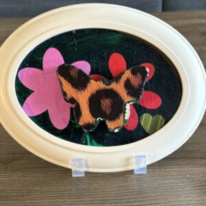 The piece is of a wood butterfly painted with orange and black patterned wings in silhouette framed in white wood. It is mounted against a dark green background with colorful shapes: a large pink flower shape on the left, a smaller red flower shape to the right, and a green heart shape below.