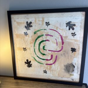 The artwork features a buttercream and gold shimmer paint background. The circular labyrinth contains curved lines in green and magenta at the center, resembling a stylized maze. Surrounding these lines are eight large and small black, simple leaf shapes scattered along the white space. The edge resembles a lace fabric design.